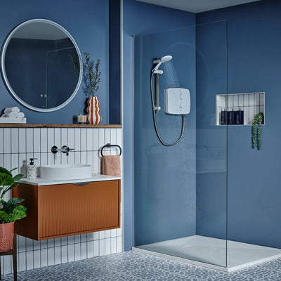 A modern bathroom setting showcasing a white Triton T90SR electric shower unit mounted on a crisp blue wall inside a glass-paneled walk-in shower tray. The surrounding space features dark blue accent walls, patterned floor tiles, a recessed shelf niche, and a wall-hung timber vanity basin unit